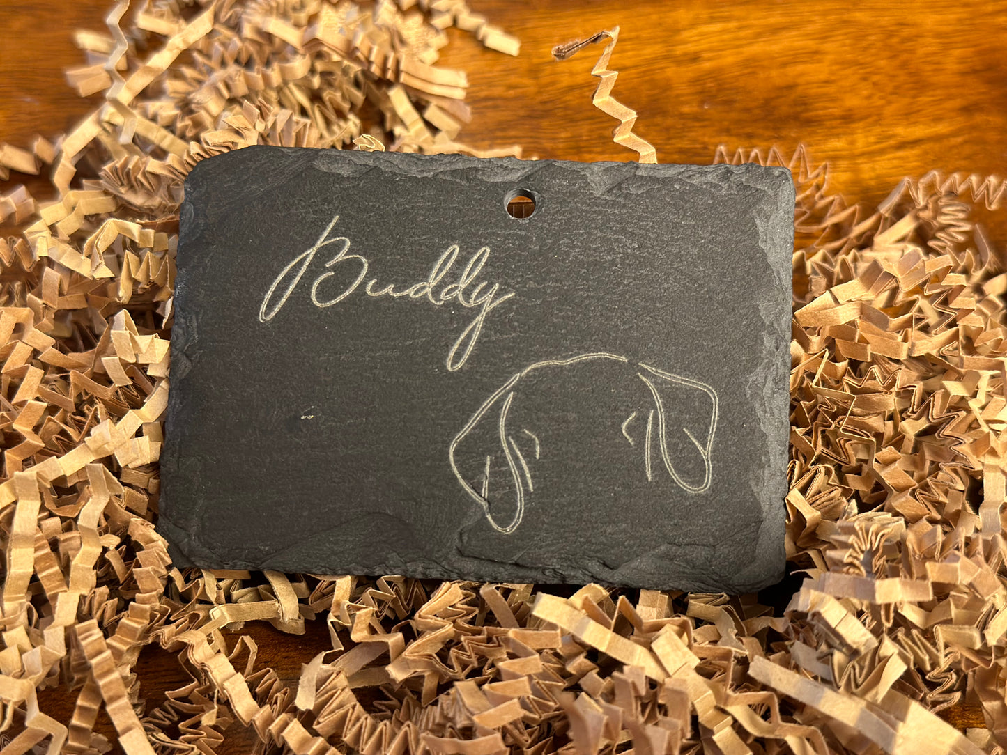 Personalized plaque made from natural slate with the name 'Buddy' and a pet silhouette, pictured on a bed of shredded brown packing material.