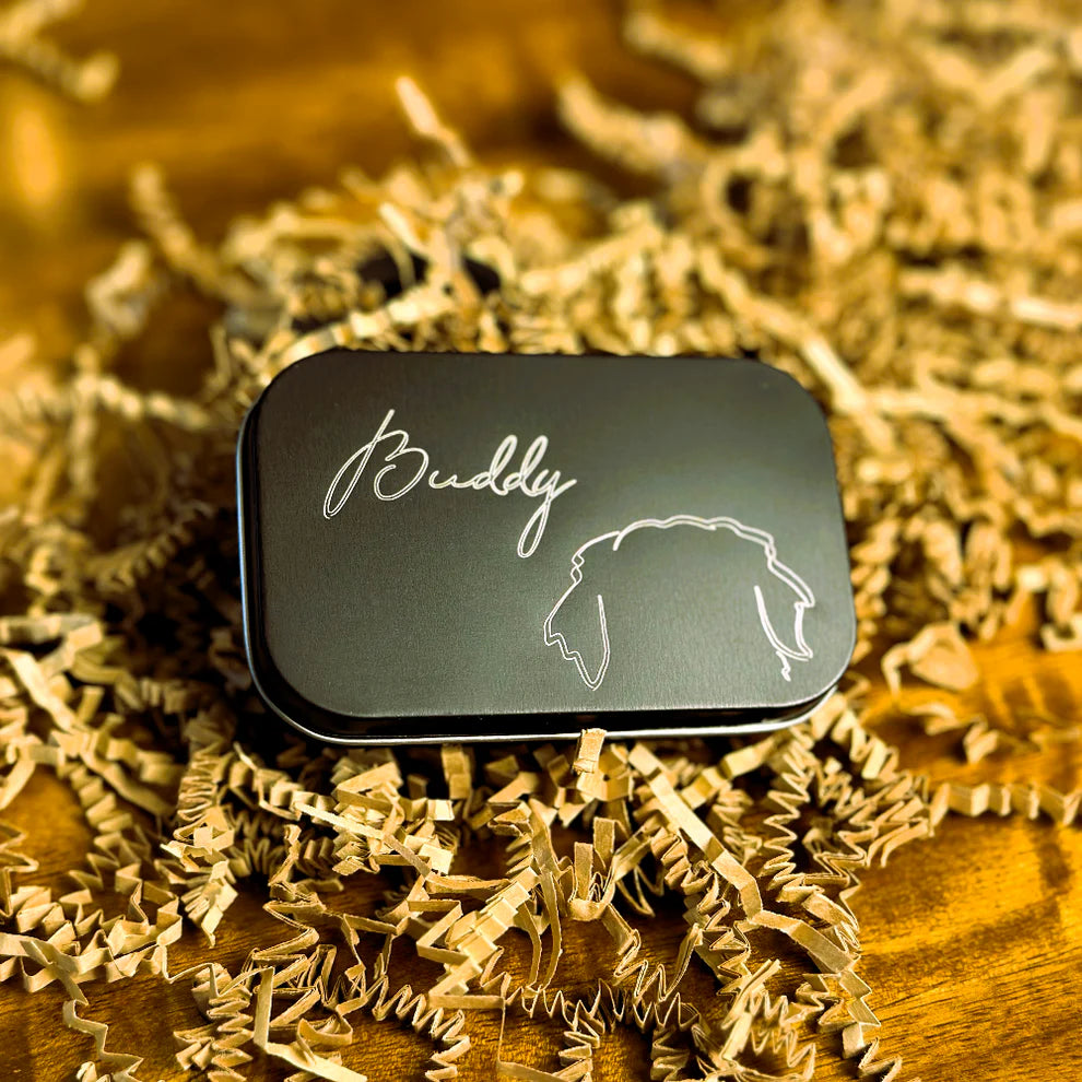 Black tin treat box engraved with 'Buddy' and a dog illustration on a bed of gold shredded paper.